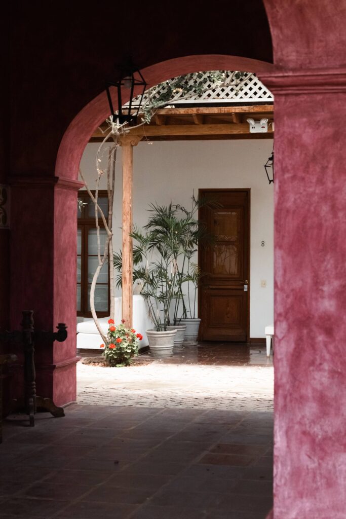 Sunlit hotel courtyard with red walls, rustic archway, potted plants, wooden door, and pergola seating area.