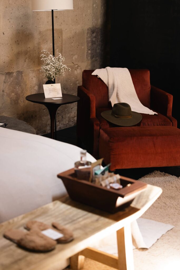 Cozy hospitality interior with red armchair, white throw, green hat, side table with flowers, and stone wall.
