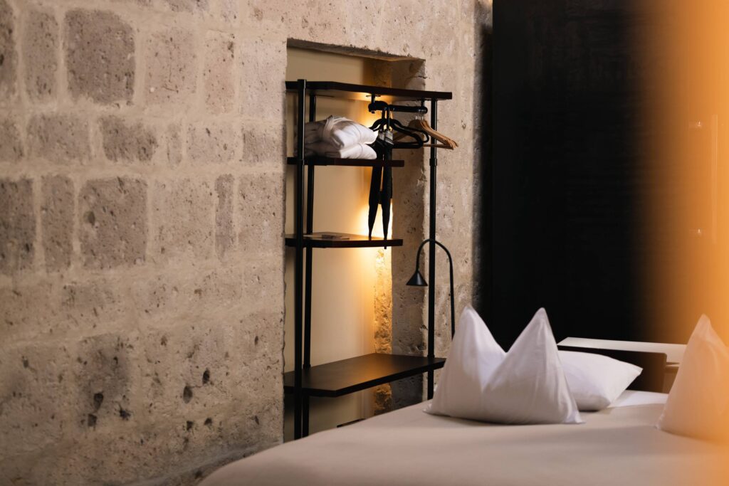 Modern bedroom corner with a textured stone wall, black metal shelves, white linens, and warm ambient lighting.