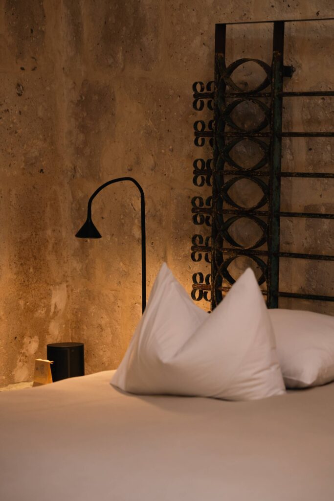 Rustic bedroom corner with crisp white pillows, a wrought iron headboard, and a textured stone wall.