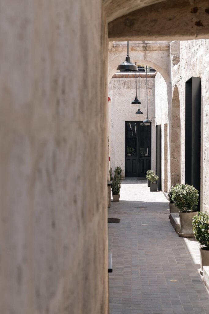 Sunlit narrow corridor with beige stone arches, black pendant lights, green plants, and a black double-door entrance.