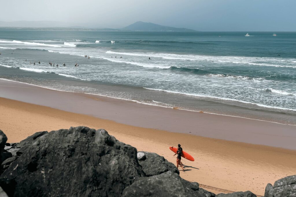 Surfer walking along a sandy beach with a red surfboard, waves, rocks, and distant mountains.