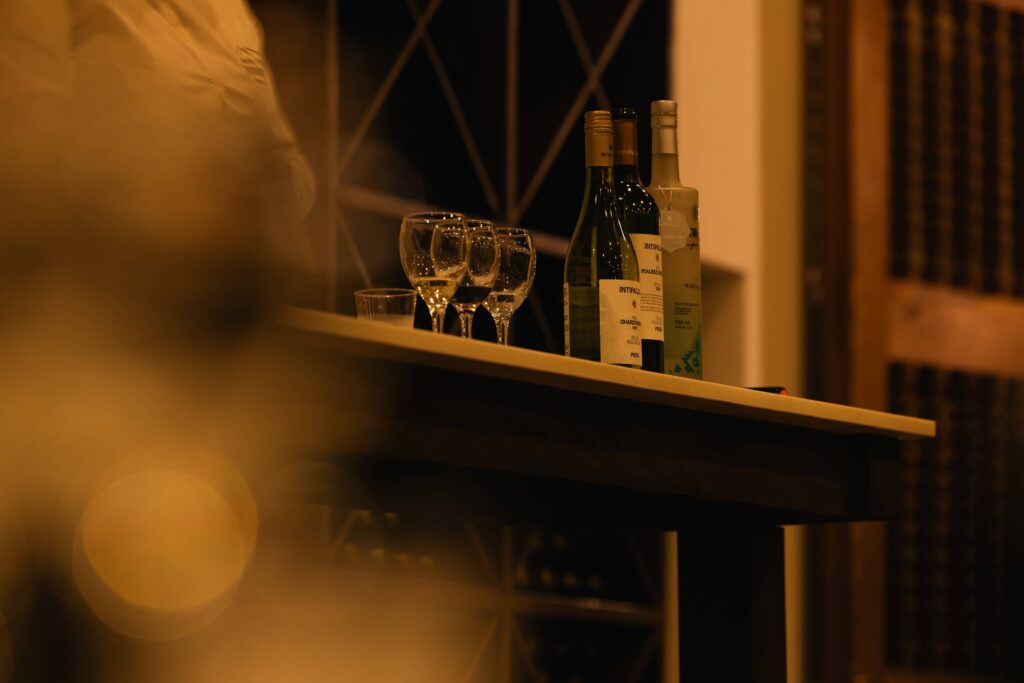 Cozy bar counter with three wine bottles and four glasses in warm, dim lighting.